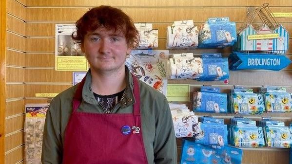 Dylan Haycock is standing in a shop surrounded by seaside-themed gifts. He has red hair and is wearing an olive green jumper and a red apron.