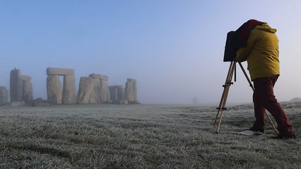 A man in a yellow coat with his head under the cloth of an old camera on a tripod, photographing Stonehenge at dusk