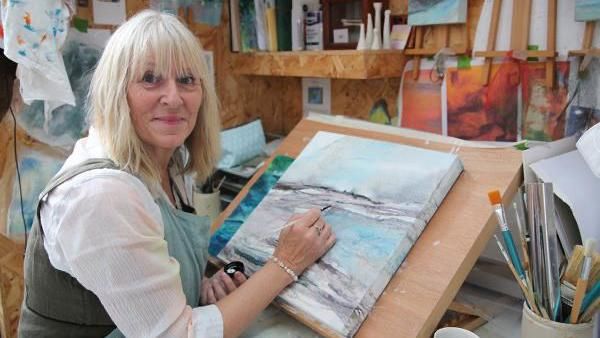 Jayne Gaze wearing a white blouse and a grey apron. She is holding a paintbrush and sitting next to a painted canvas