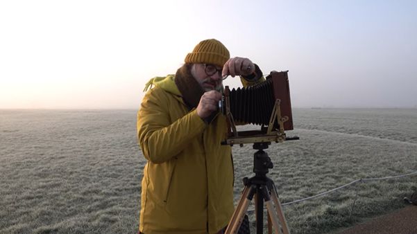 Frank Menger, in a yellow coat and hat, adjusting a camera from the 1890s outside