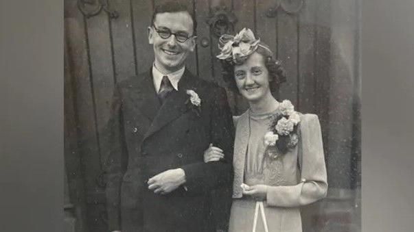 John and Blodwen Tinniswood on their wedding day, with him in glasses and a double-breasted dark suit and her with a fascinator and a pale two-piece suit, holding a silver horseshoe