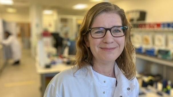 A woman smiling at the camera, wearing glasses infront of a laboratory