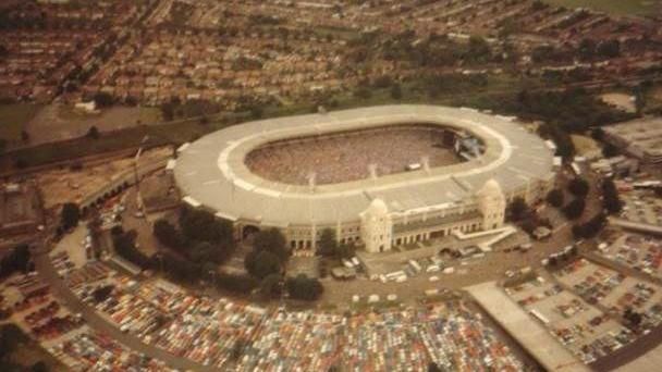 Wembley Stadium on 13 July, 1985, on the morning of Live Aid. The stadium is full, with hundreds of cars parked outside