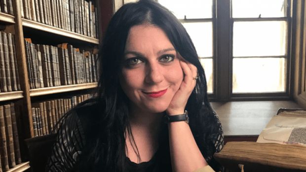 Professor Janina Ramirez sitting at a desk in an old library. She has her chin resting on her hand and is looking at the camera and smiling, wearing all black with red lipstick.