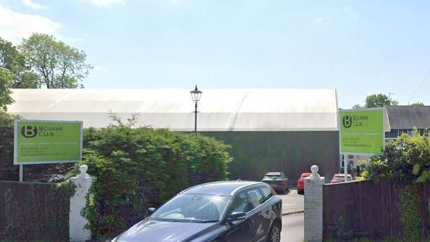 The entrance to the Bourne Tennis Club with green signs and the name of the club as well as a road sign which reads Frensham Road