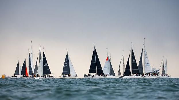 A group of sailing boats on the water with a slight ripple in the water and clear skies behind the boats they all have a mix of dark blue and white sails