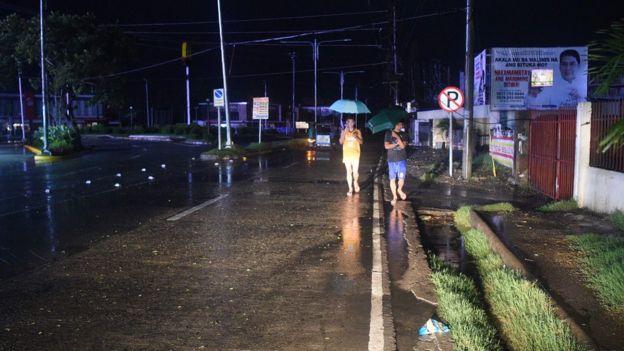 Pedestres caminham em rua deserta na cidade de Tuguegarao durante noite e segurando guardas-chuvas