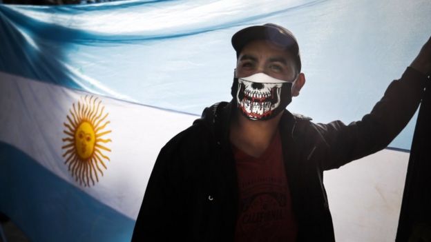 El hombre usa una máscara con un diseño de cráneo sangrante, con la bandera argentina en el fondo