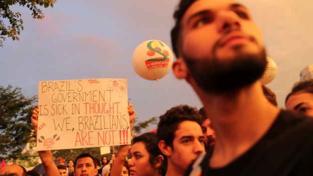 Rostos de manifestantes em São Paulo, entre os quais um que segura placa dizendo em inglês: 'O governo brasileiro é doente no pensamento; nós, brasileiros, não'