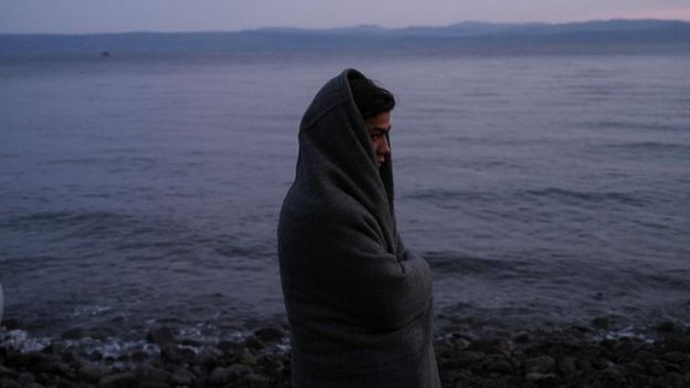A migrant covered with a blanket stands on a beach on Lesbos