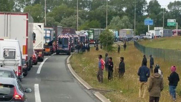 Migrants standing by the side of a road with traffic lined up