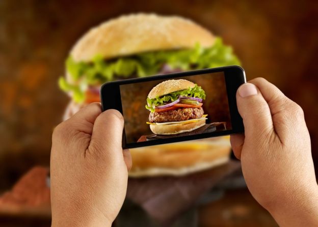 Hombre tomándole una foto con su celular a una hamburguesa.