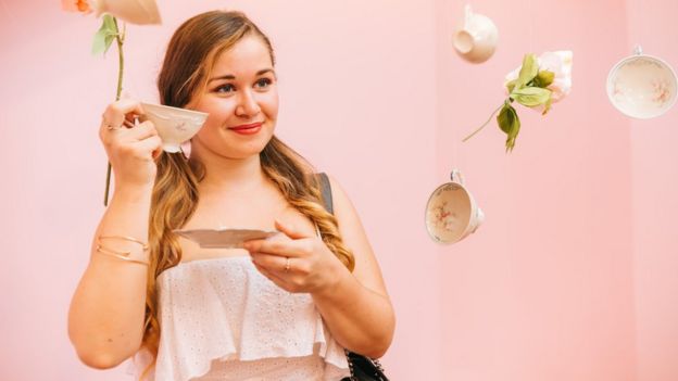 A woman holds a cup in front of a pink background