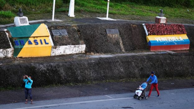 Famílias caminham em estrada em cujas margens estão duas pinturas com dizeres e cores respectivas de cada país: Brasil e Venezuela