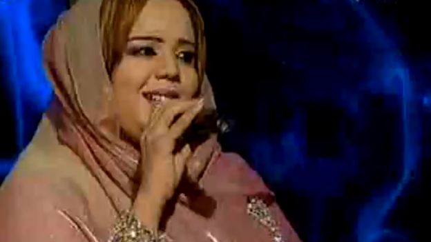 Nada Algalaa sings into a microphone as she performs on stage, wearing a pale pink dress and headscarf.