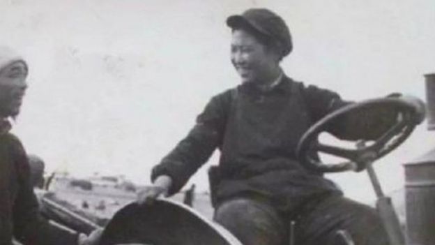 Foto em preto e branco de Liang Jun jovem sorrindo de cima de trator, observada por outra pessoa