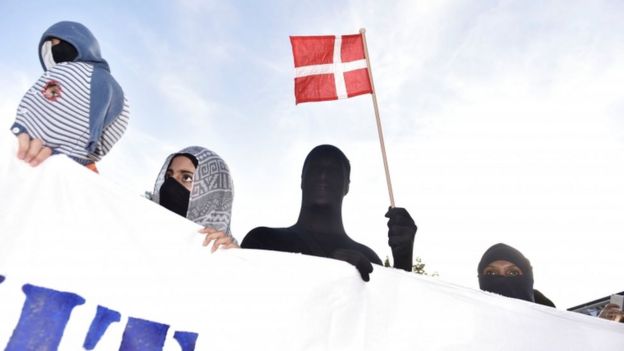 Imagem mostra pessoas com o rosto coberto e um boneco empunhando a bandeira da Dinamarca, em protesto realizado na cidade de Copenhague, no primeiro dia da implementação da proibição do véu no país