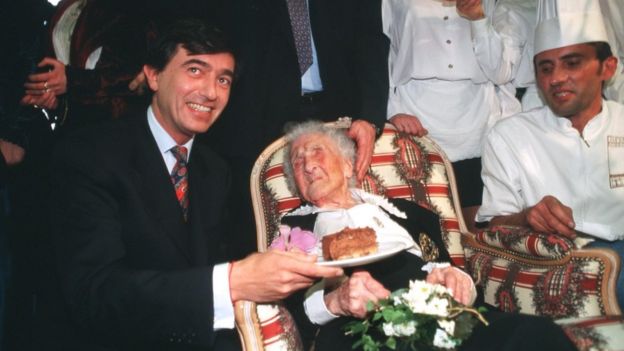 Calment na cadeira em seu 120º aniversário, ao lado de um homem sorrindo e segurando uma fatia de bolo