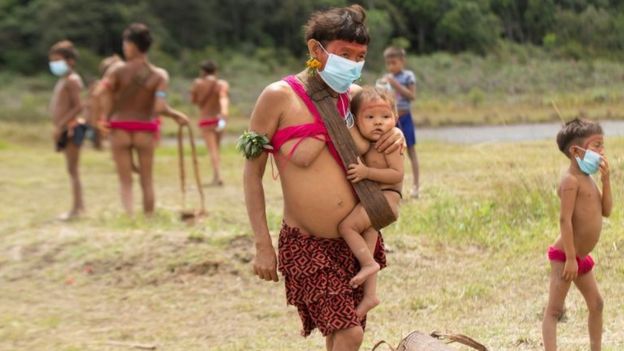 Indígena com bebê no colo, rodeado por vários outros espalhados em campo