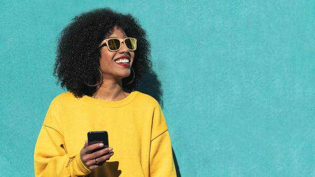 Mulher com cabelo black, óculos escuros, moletom amarelo e celular na mão - o uso de celular gera milhares de dados pessoais, nas mãos de empresas