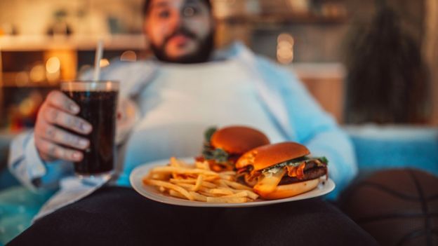 Homem sentado em um sofá degustando uma comida rica em gorduras.