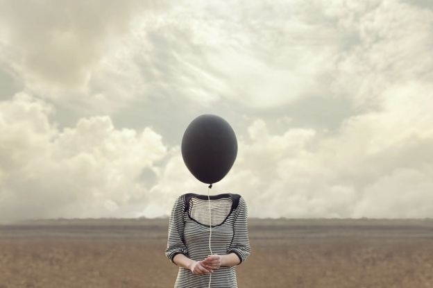 Photo of a woman on an open field, but the woman's head has been replaced by a black balloon, and the feeling is eery