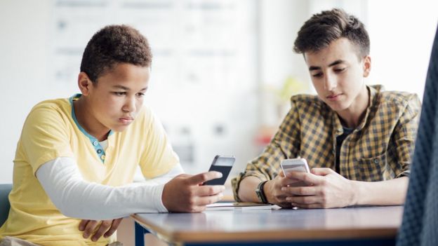 Dois adolescentes sentados à mesa olham para seus celulares