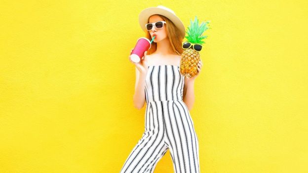 Modèle féminin avec un ananas et une tasse de jus. Elle porte un pantalon rayé blanc et un chapeau rond. Elle est debout sur fond jaune