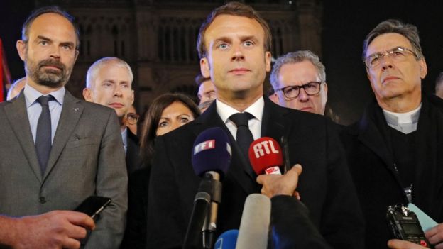 Macron perto de microfones e rodeado por assessores e membros da Igreja Católica nos arredores da Notre-Dame, durante noite