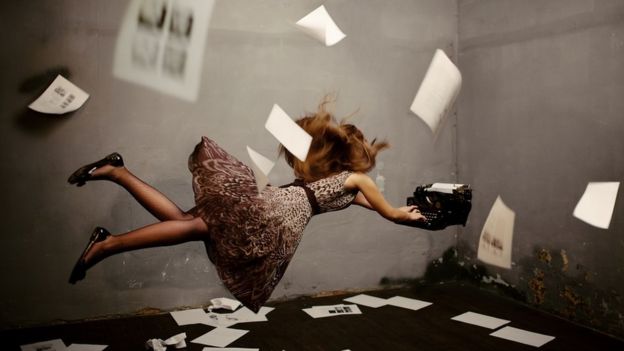 Surreal concept image: a young woman writing, suspended in mid-air, typing on a flying typewriter. There are pages on the floor and floating in the air around her.