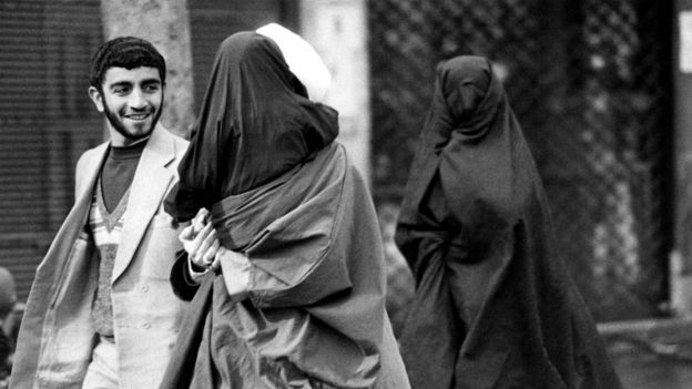 ملا يرتدي عمامة بيضاء وهو يحمل طفلته المرتدية شادورا أسود ويتكلم إلى رجل، وراءه زوجته مرتدية أيضا شادورا أسود ووجهها مغطى - شباط/فبراير 1980
