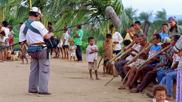 Cena da gravação do documentário 'No Caminho da Expedição Langsdorf', em uma comunidade indígena - homens estão sentados tocando um instrumento de sopro longo