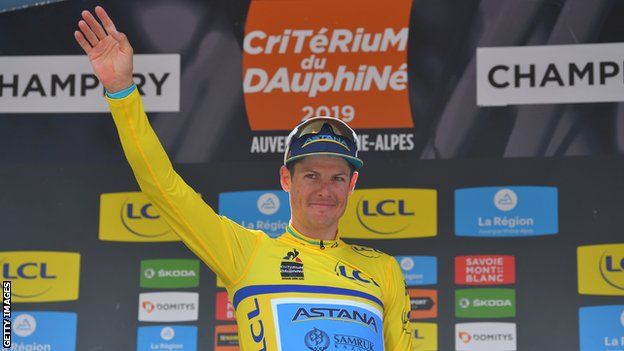 Astana rider Jakob Fuglsang waves on the podium at the 2019 Criterium du Dauphine while wearing the leader's yellow jersey
