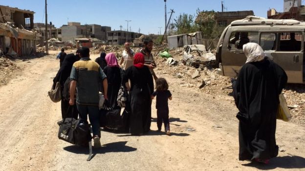 A family group walks along a road lined with burnt out cars and destroyed buildings