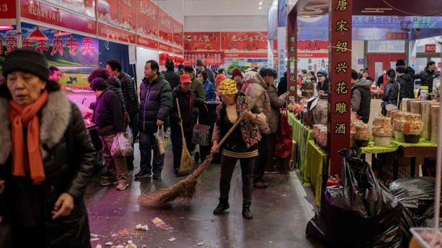 Corredor de mercado em Pequim, onde se ver mulher varrendo o chão e diversos consumidores caminhando entre estandes com sacolas