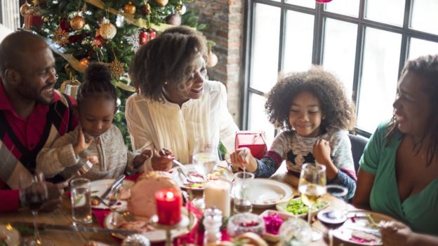Família come ceia de Natal ao redor de uma mesa, com árvore decorada ao fundo