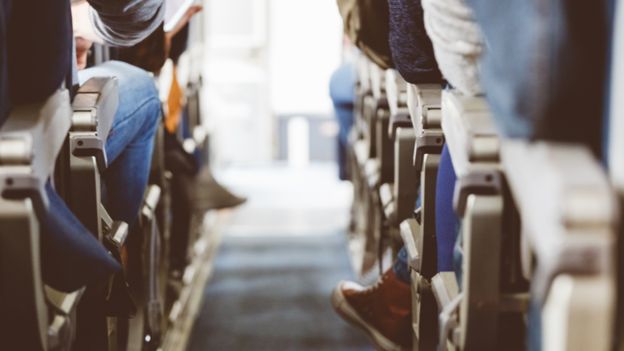 Imagem mostra corredor de avião, exibindo passageiros em seus assentos