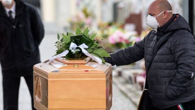 Homem de máscara põe a mão sobre caixão durante um velório na Itália