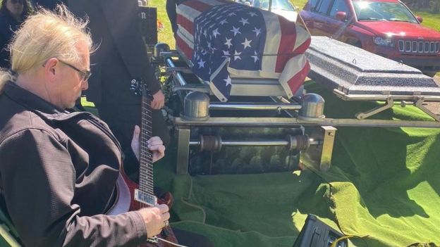 Homem tocando guitarra ao lado de caixão do pastor Spradlin, coberto por bandeira dos Estados Unidos sobre grama