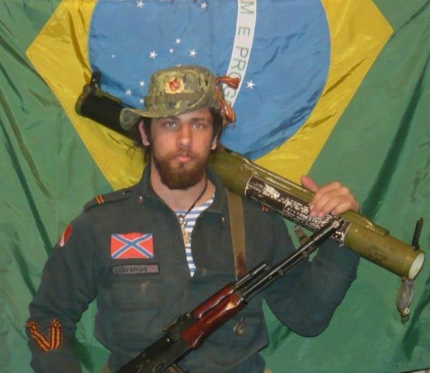 Rafael Lusvarghi com armamento pesado em frente à bandeira brasileira