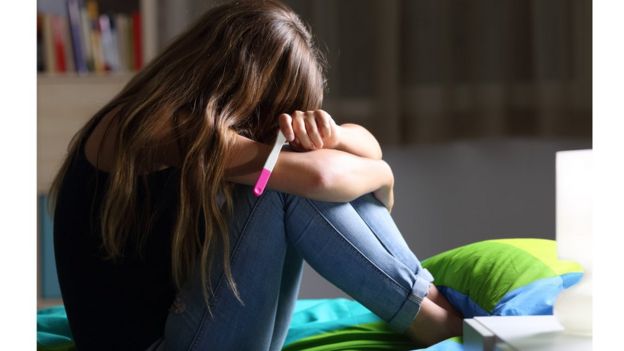 Adolescente sentada na cama com teste de gravidez em uma das mãos