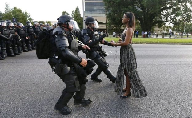 Fotógrafo de Nova Orleans registrou imagem de uma jovem negra de vestido, de pé e aparentando calma diante do que parece ser a chegada esbaforida de dois policiais armados e trajando equipamento completo de choque. A foto viralizou internacionalmente.