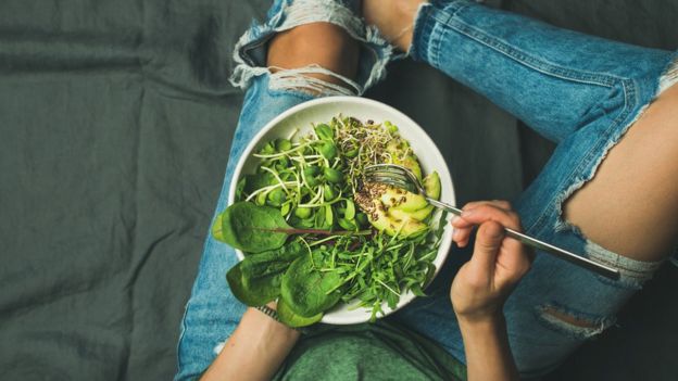 Young woman sitting holding a bowl full of spinach, rocket and avocado.