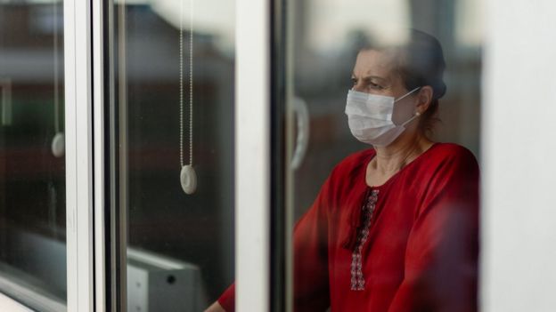 Una mujer con mascarilla parada detrás de en una ventana.