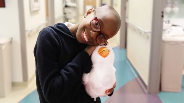Menina em tratamento para câncer, careca e sorridente, de óculos, abraça seu pato-robô