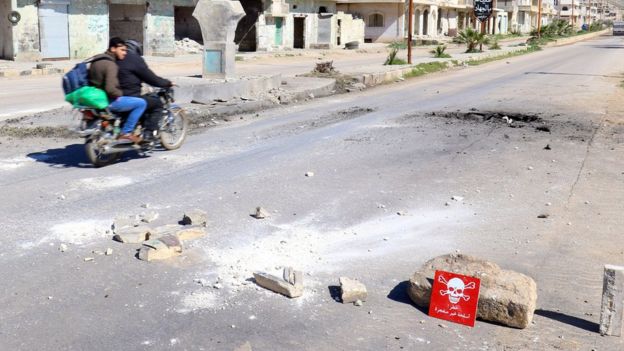 Men ride a motorbike past a hazard sign at a site hit in an alleged chemical weapons attack in Khan Sheikhoun, Syria (5 April 2017)