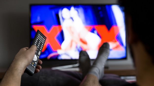 Imagem desfocada na televisão mostra duas mulheres, indicando conteúdo pornográfico, assistido por homem com controle remoto