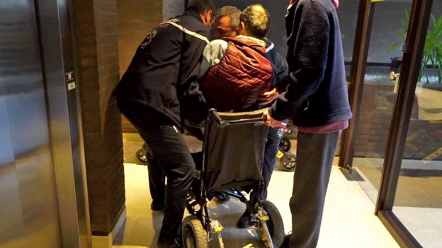 Alain sendo transferido de sua cadeira de rodas, com a ajuda de três homens. Homem cogitava fazer eutanásia, mas morreu antes de complicações rspiratórias