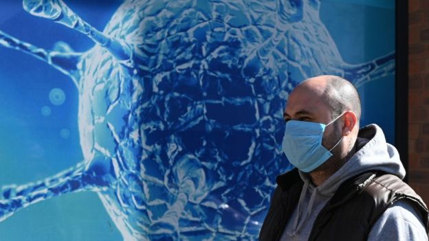 Um homem com uma máscara passa na frente de uma parede com uma ilustração do coronavírus