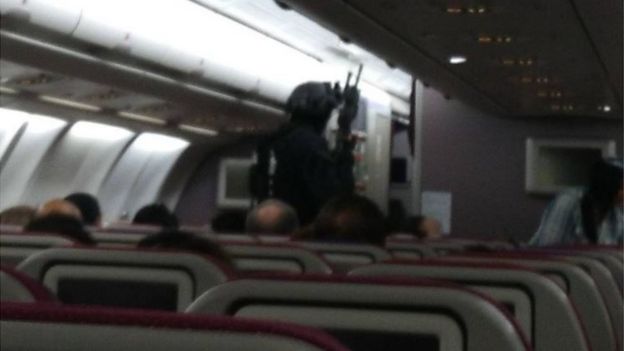 A blurry photo shows passengers seated on the plane, while a heavily armed man in full tactical gear and what appears to be a rifle walks towards the front of the plane